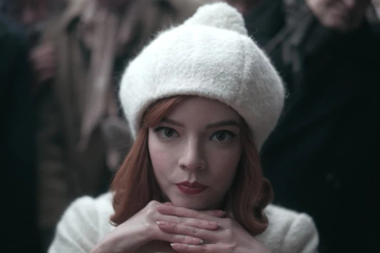 Beth Harmon, wearing white, rests her chin on her hands as she contemplates a move.