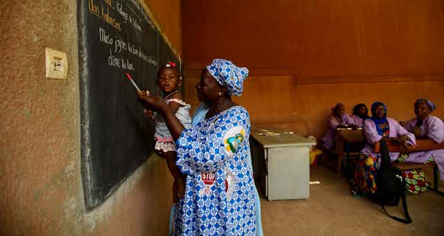 A woman holds a baby in front of a blackboard.