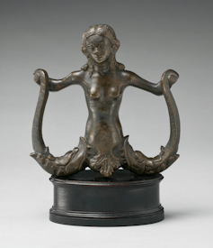 A bronze statue of a mermaid with two tails. She is holding a tail in each hand.