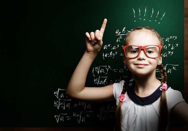 Young girl pointing up in front of blackboard.