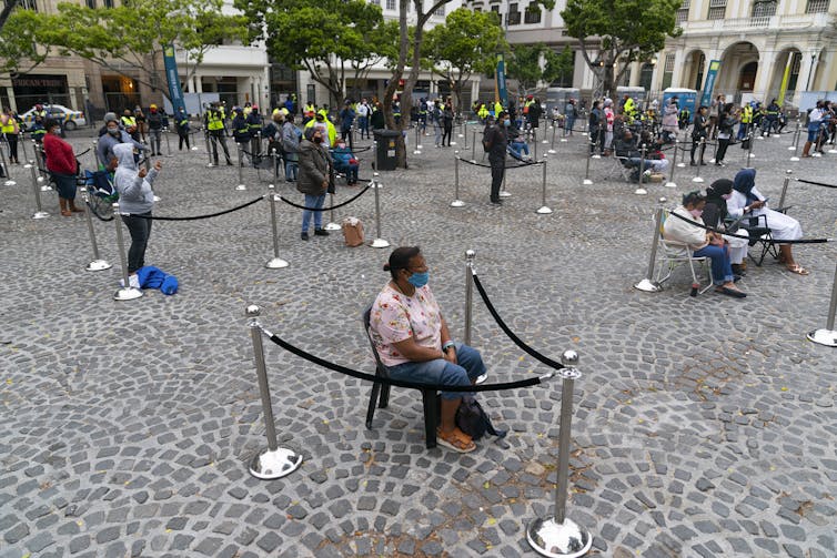 In a village square, people sit wearing masks at a distance from one another, their seating area marketed by ropes.
