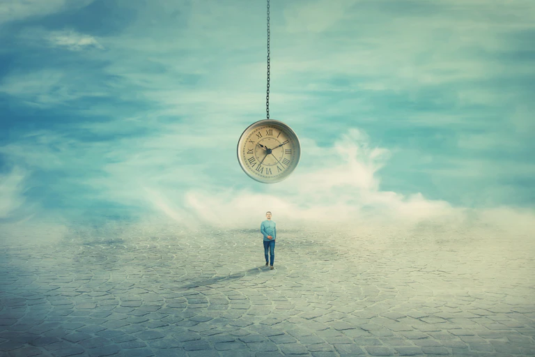 A man stands beneath a hanging clock on a cloudy street.