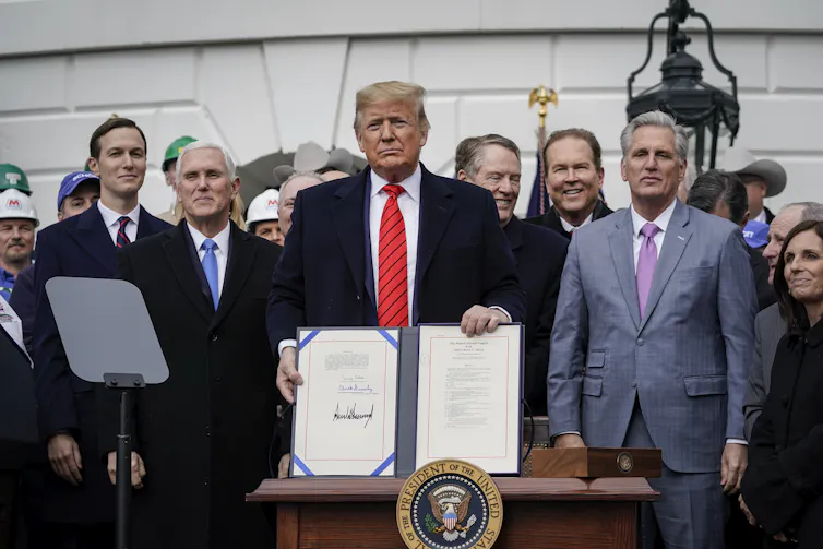 Trump stands with a document in front of him, surrounded by people including Vice President Mike Pense and son-in-law Jared Kushner