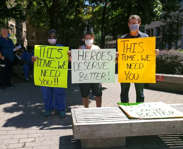 Three people wearing face masks hold hand-lettered signs asking for public support of health-care workers.