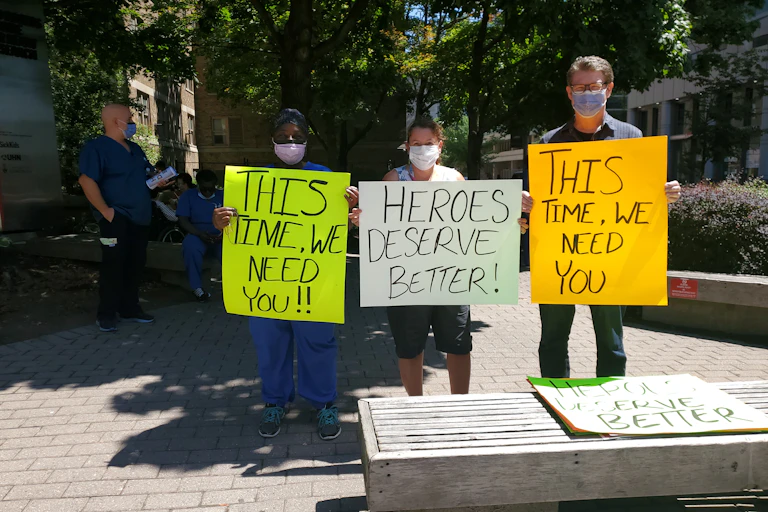 Three people wearing face masks hold hand-lettered signs asking for public support of health-care workers.