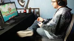 A young man sits in front of a computer with a game console.