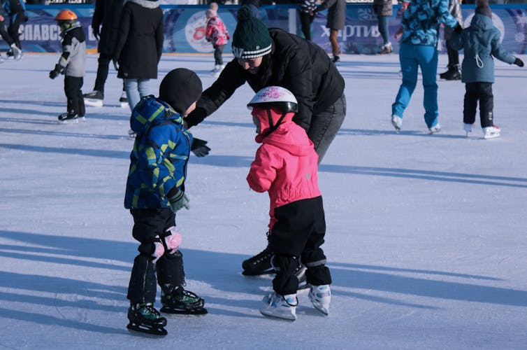An adult bends down to talk to children; all are on ice skates on an outdoor ice rink.