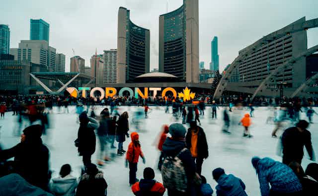People skate on an outdoor ice rink. City Hall and Toronto sign are in the background. 