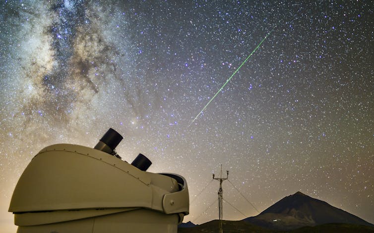 An asteroid streak across the sky with a volcano and telescope in the foreground.