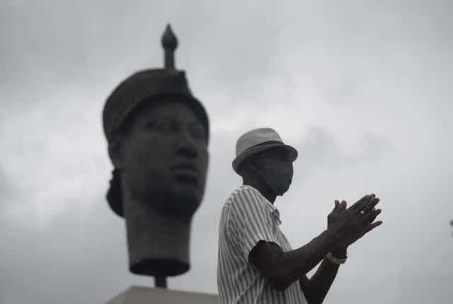 In black and white, the image shows a man in profile in a casual hat, his hands held together in front of him. Behind him is a large statue of a traditional African face.  