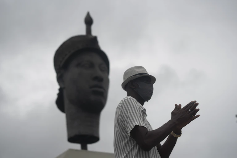 In black and white, the image shows a man in profile in a casual hat, his hands held together in front of him. Behind him is a large statue of a traditional African face.
