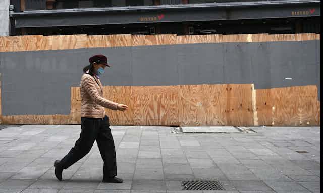 Person wearing a mask walking past a boarded up bistro.