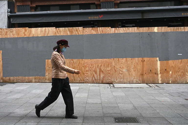 Person wearing a mask walking past a boarded up bistro.