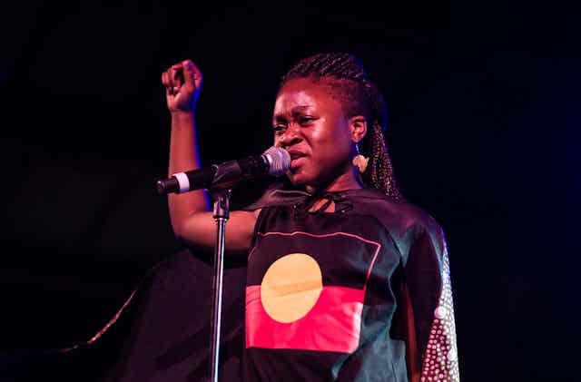A young woman stands talking into a microphone, her right arm raised in a fist, wearing a black cape and a blouse with a large yellow circle on it.