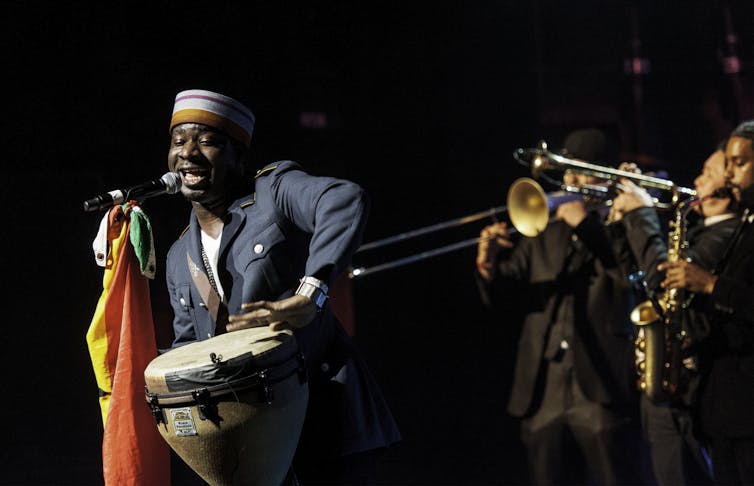 A man plays African drums and sings into a microphone, behind him a row of trumpeters and saxophonists.