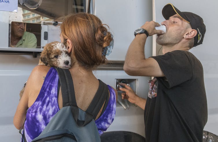 Woman with a dog waits at a white van while a man drinks from a tiny cup