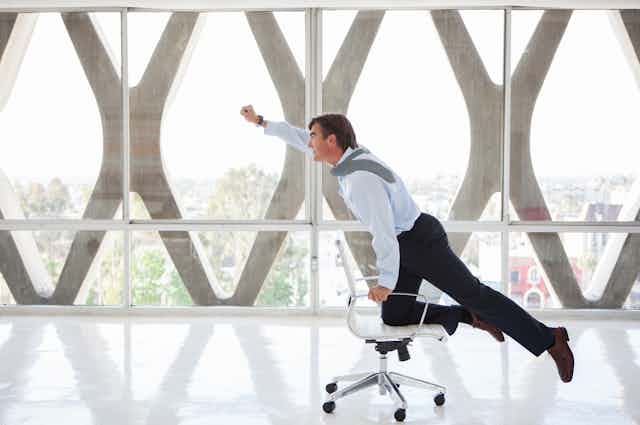 Un hombre de negocios atraviesa una sala vacía sobre una silla con ruedas imitando a Supermán.