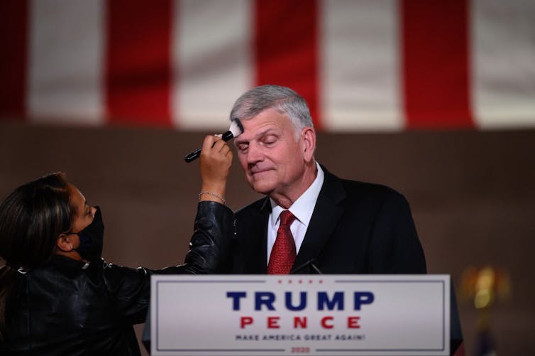 A woman brushes makeup on Franklin Graham's forehead as he stands at a podium during the 2020 Republican National Convention.