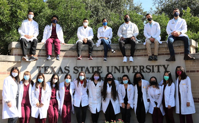 group of students in white medical coats and face masks gather around texas state university sign