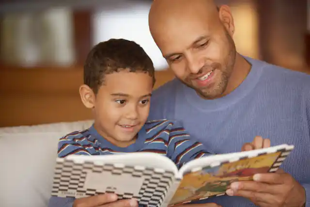 Father and son reading book while sitting on couch.