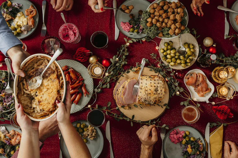 Someone passes a dish filled with food to someone else amid a table that displays many types of holiday foods.