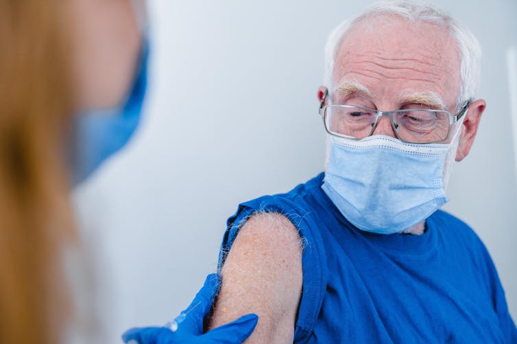 An older man wearing a mask has his sleeve rolled up in preparation for a vaccination.