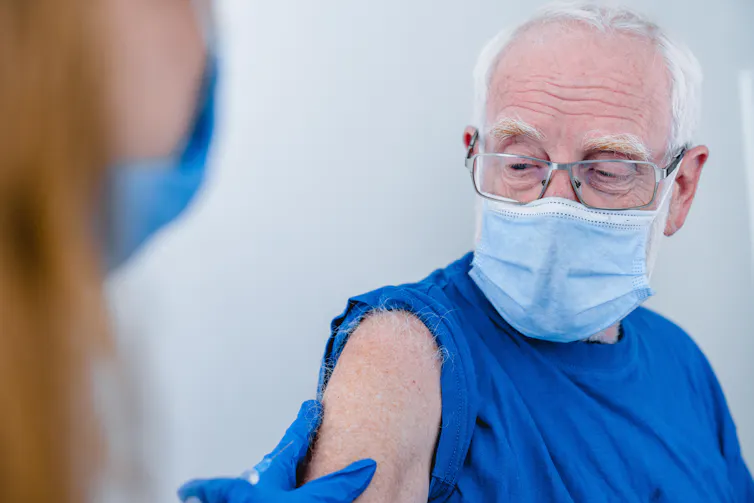 An older man wearing a mask has his sleeve rolled up in preparation for a vaccination.