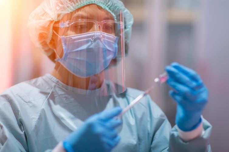 A health worker dressed in full PPE holds a syringe and a vial.