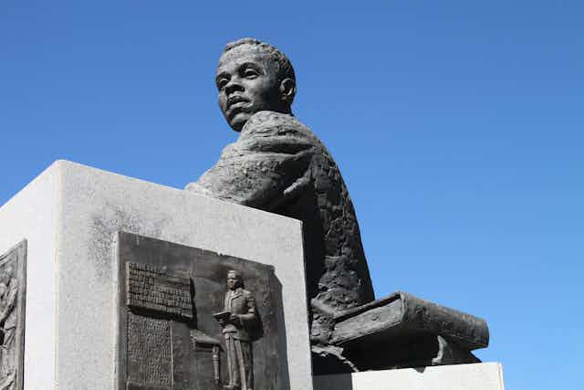 A sculpture of a young man in a jacket, seated with a book next to him. He's gazing towards the horizon, blue sky behind him.