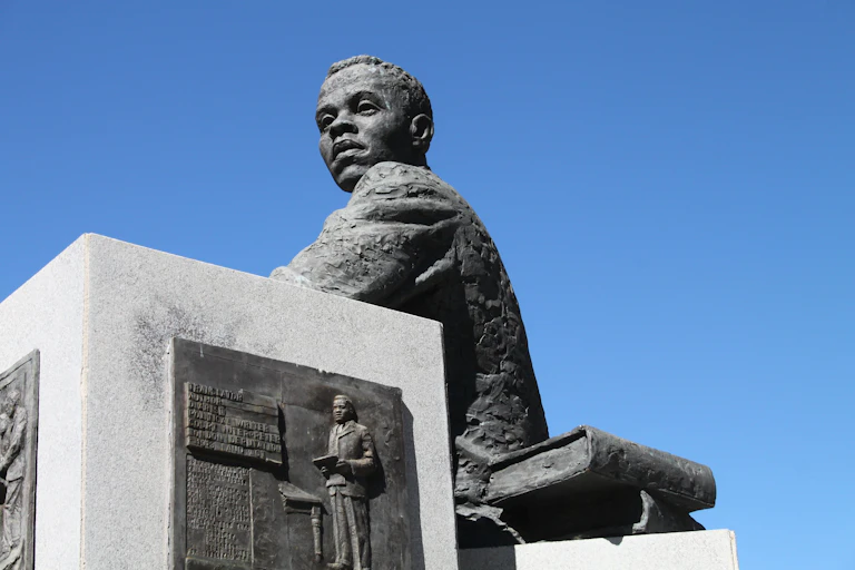 A sculpture of a young man in a jacket, seated with a book next to him. He's gazing towards the horizon, blue sky behind him.