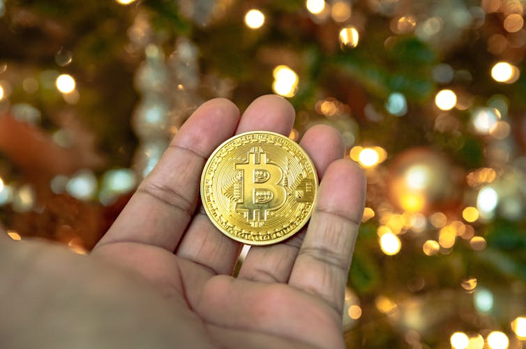 A person holds a gold bitcoin coin in front of a Christmas tree