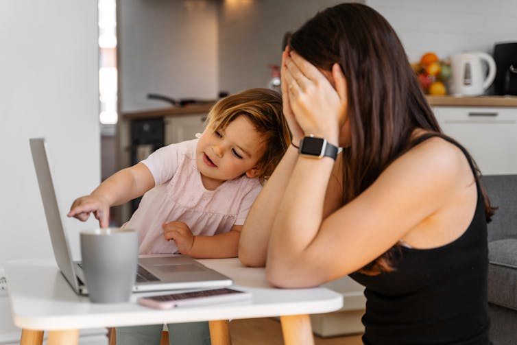 A working mum holds her head in her hands as her toddler interferes with her laptop.