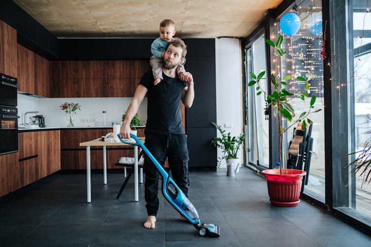 A dad vacuums with a toddler on his shoulders.