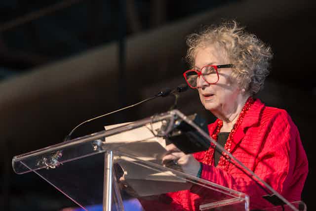 Margaret Atwood stands at an acrylic podium, in a red blazer and red-framed glasses