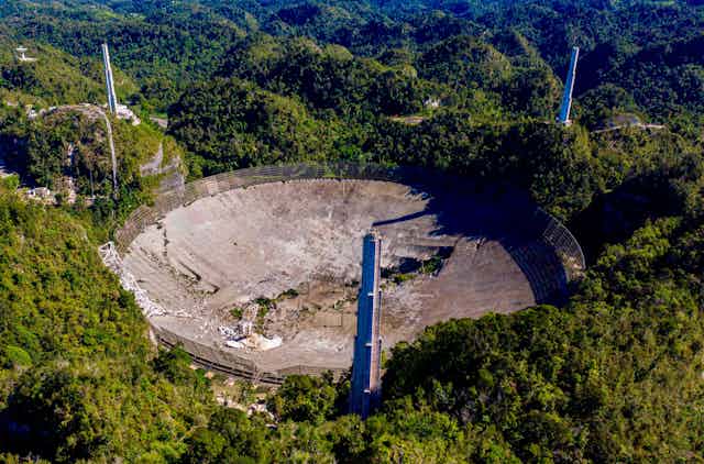 An arial view of an enormous crumbling telescope dish surrounded by hilly terrain.