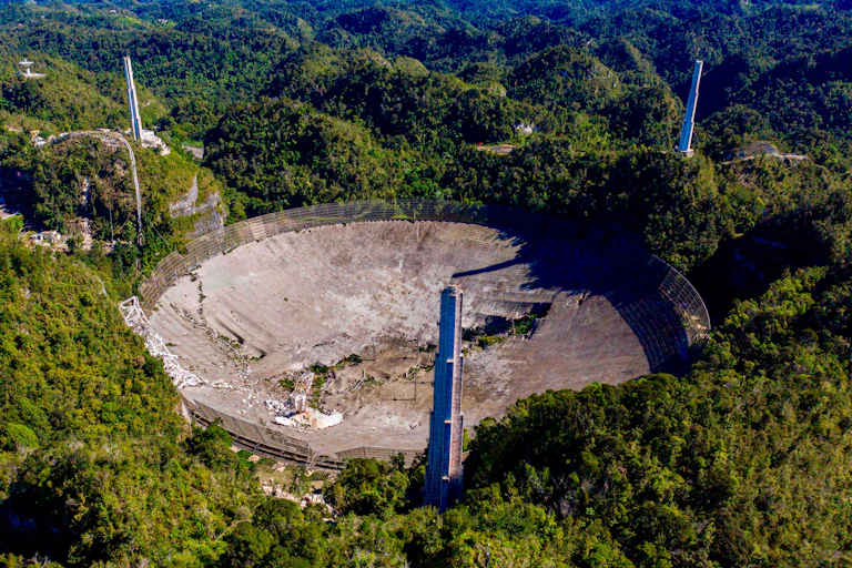 An arial view of an enormous crumbling telescope dish surrounded by hilly terrain.