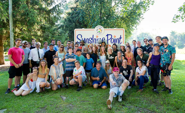 (A crowd outside in front of 'Sunshine Point' sign.