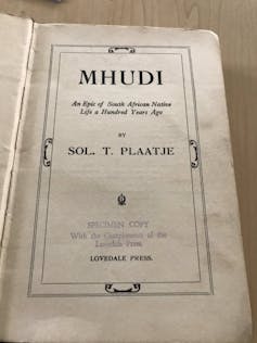 An old book open at the first page, with text in an illustrated frame reading, 'Mhudi - an epic of South African Native Life a Hundred Years Ago.'