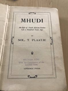 An old book open at the first page, with text in an illustrated frame reading, 'Mhudi - an epic of South African Native Life a Hundred Years Ago.'