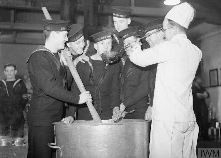 A group of sailors in uniform stirring a large cooking pot with a chef.