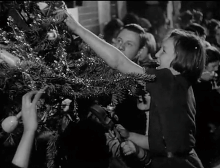 Black and white photo of a child decorating a Christmas tree with an adult in the backgrond.