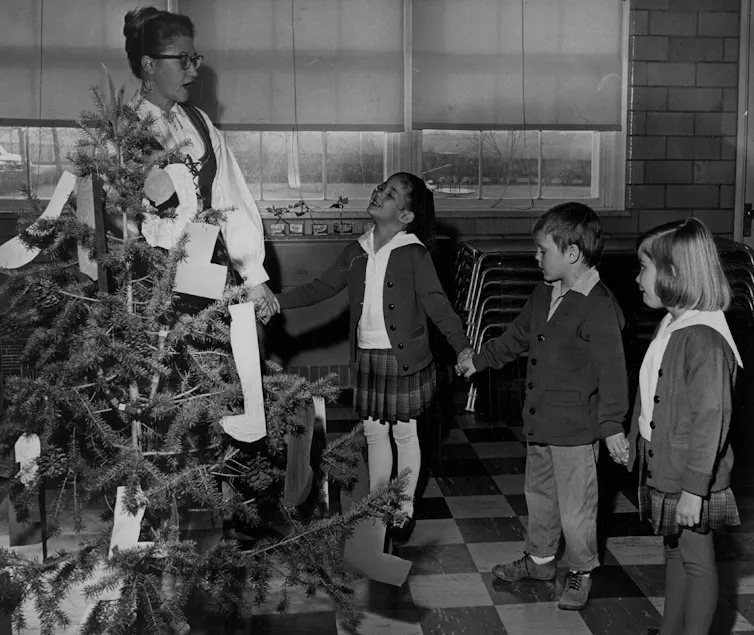 A teacher and students celebrate Christmas around a tree in school in this black-and-white photo from 1964