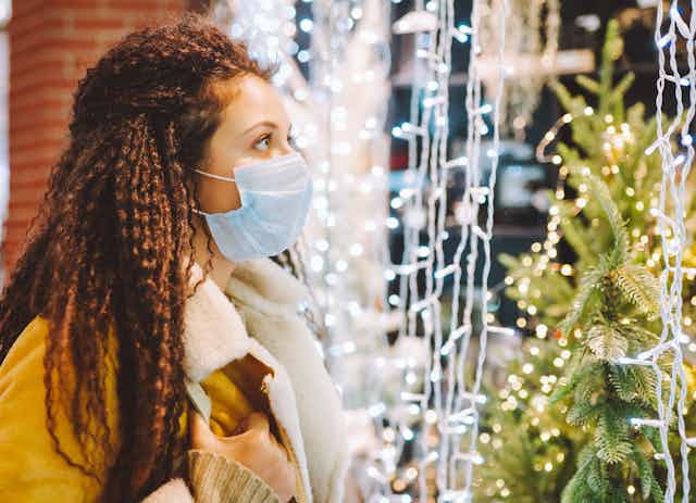 A woman in a face mask looking at a Christmas tree and Christmas lights