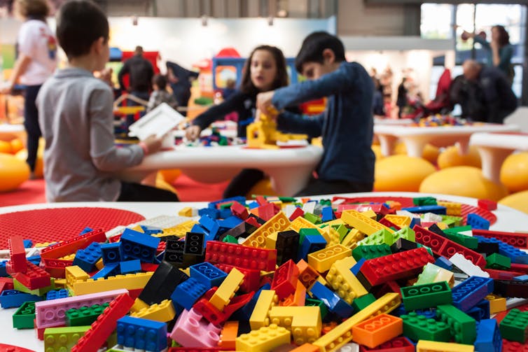 A pile of Lego bricks in the foreground with children playing with them in the background.