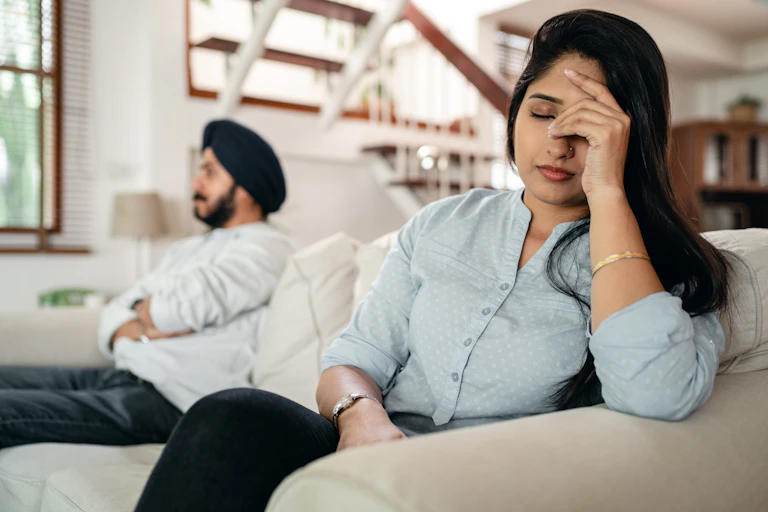 A woman at one end of a sofa looking upset, while a man sits at the other end with his arms crossed.