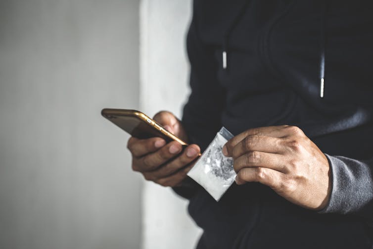 A man holds a small packet with white powder in one hand, and his phone in the other hand.