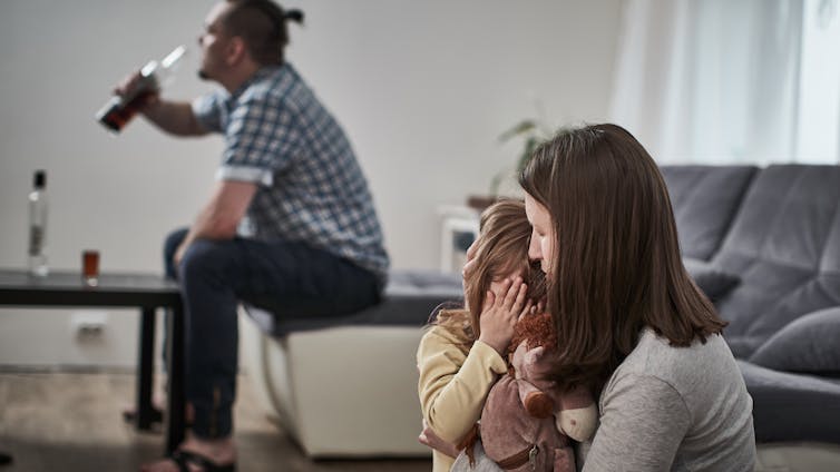 A man drinks while his wife and child sit in the room.