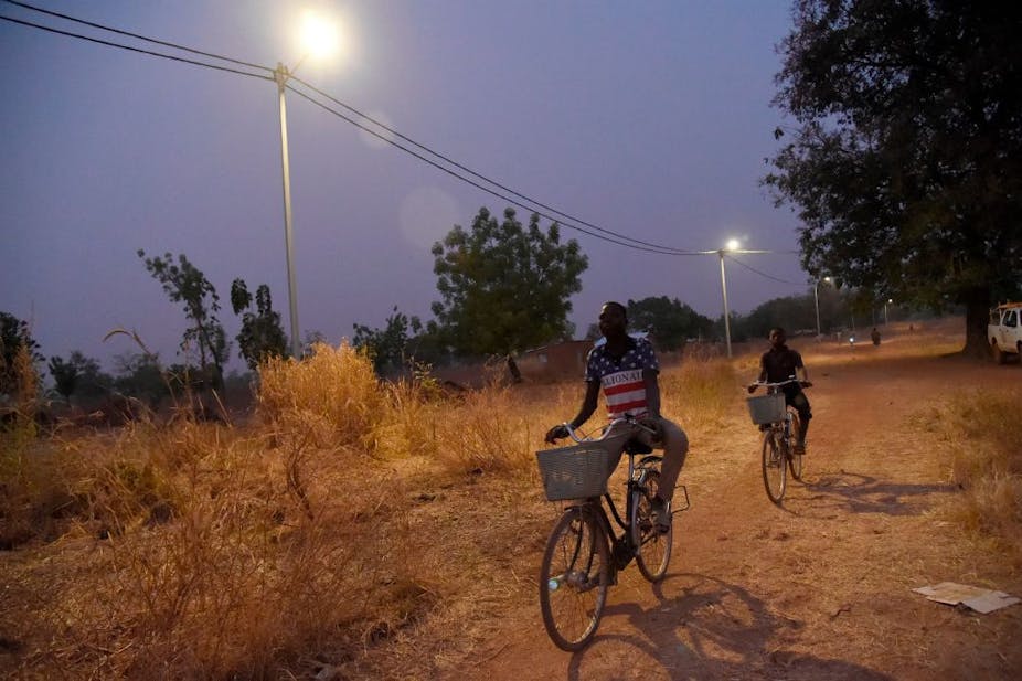 Two cyclists drive past street light poles on a dusty village pathway.