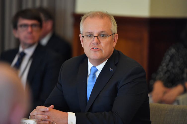 Scott Morrison sits at a table in a suit delivering remarks.