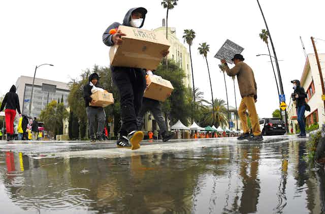 People cross a flooded street with palm trees holding cardboard boxes.